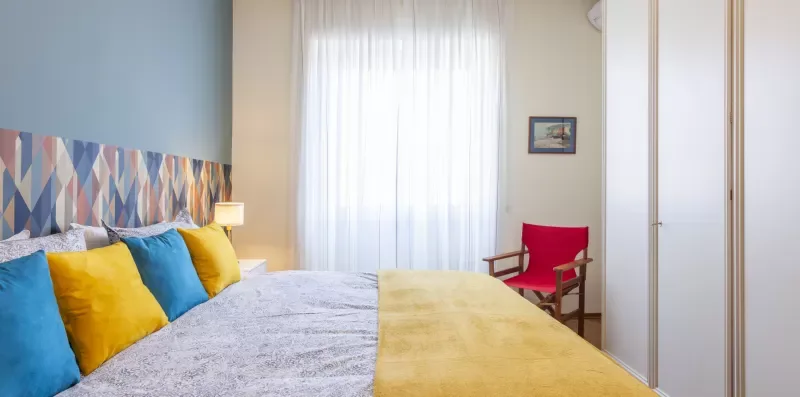 Dormitorio con una cama, cojines coloridos y una silla roja junto a una ventana luminosa.