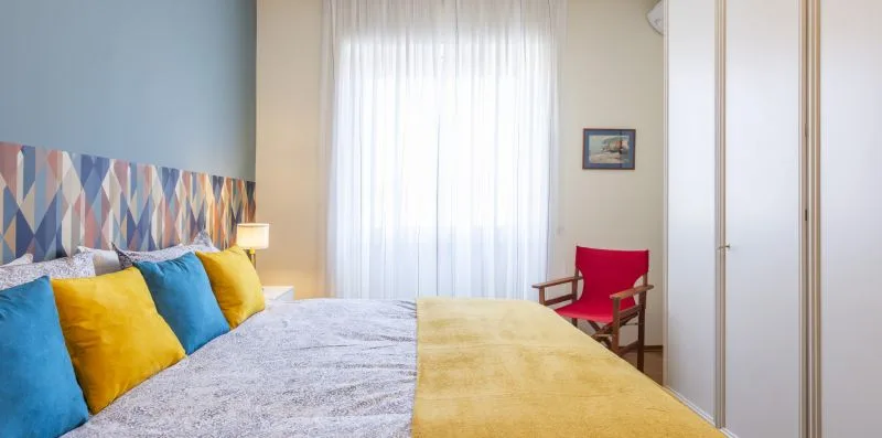 Un dormitorio con cojines coloridos sobre la cama y una silla roja junto a la ventana.
