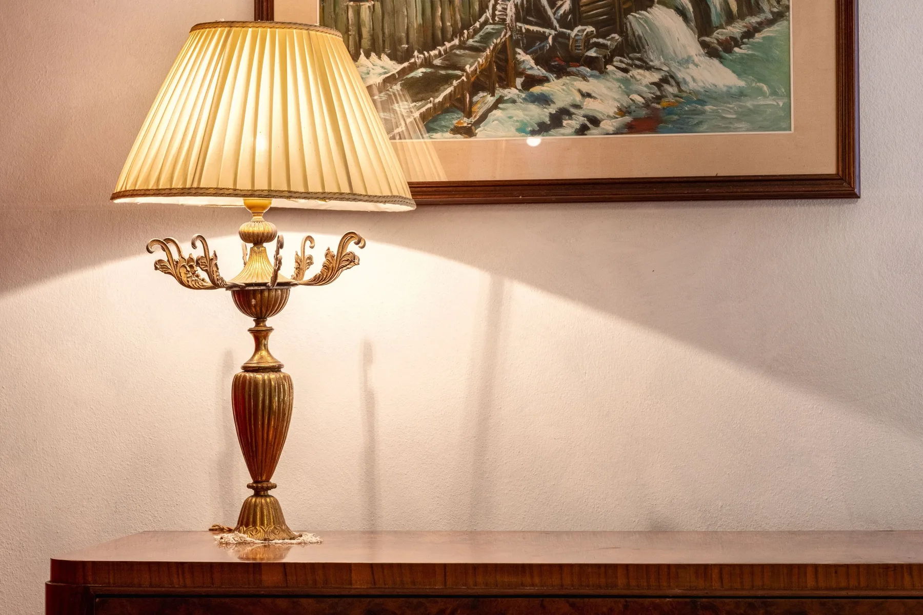 A lit table lamp stands on a wooden console beneath a framed painting.