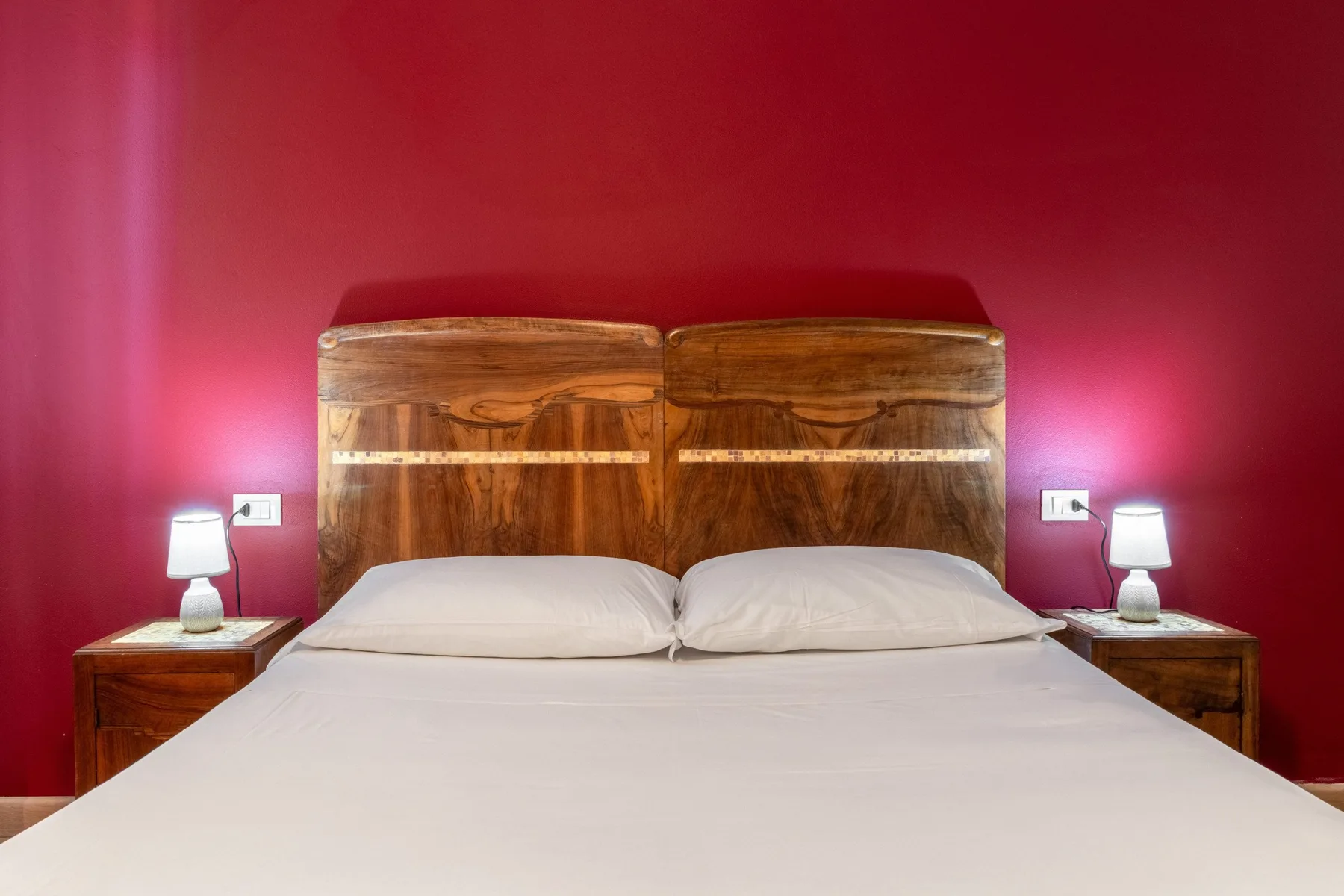 A bedroom with a bed, wooden headboard, and bedside lamps against a red wall.