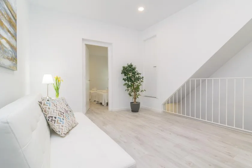 Una luminosa sala de estar tiene asientos blancos, una planta en maceta y una escalera.