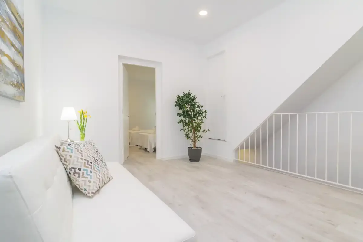 Una luminosa sala de estar con asientos blancos, una planta en maceta y una escalera.