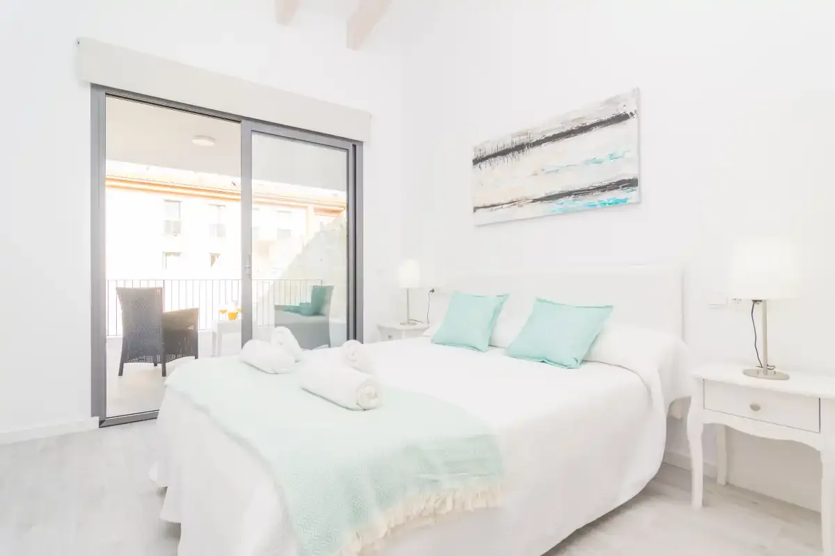 Un dormitorio luminoso con una cama blanca y puertas de cristal que dan a un balcón.