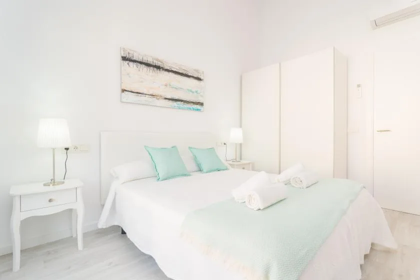 Dormitorio luminoso con cama blanca, ropa de cama verde agua y mesitas de noche.