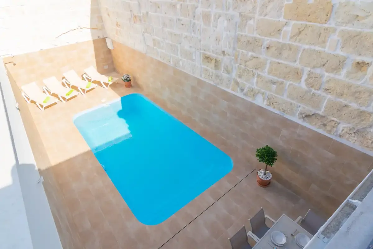 Una pequeña piscina en un patio está junto a un muro de piedra y tumbonas.