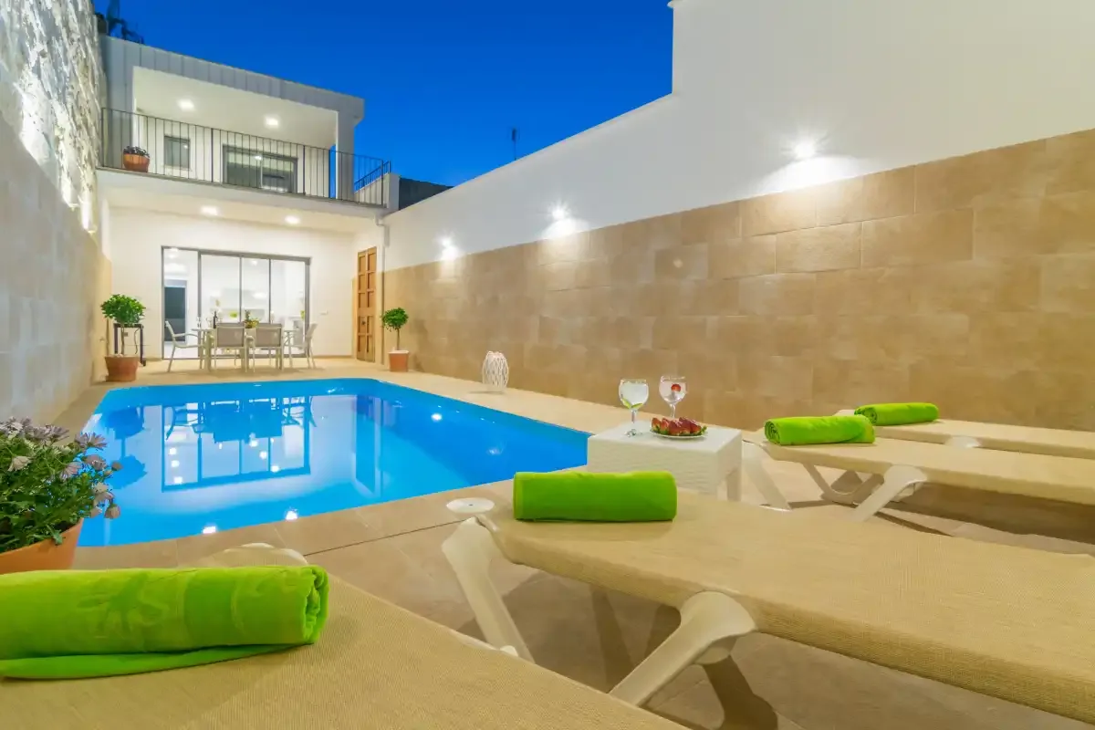 Una piscina junto a un patio de baldosas con tumbonas acolchadas y una zona de comedor.