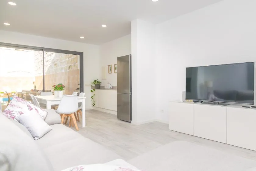 Una luminosa sala de estar y comedor con un televisor y una mesa junto a grandes ventanas.