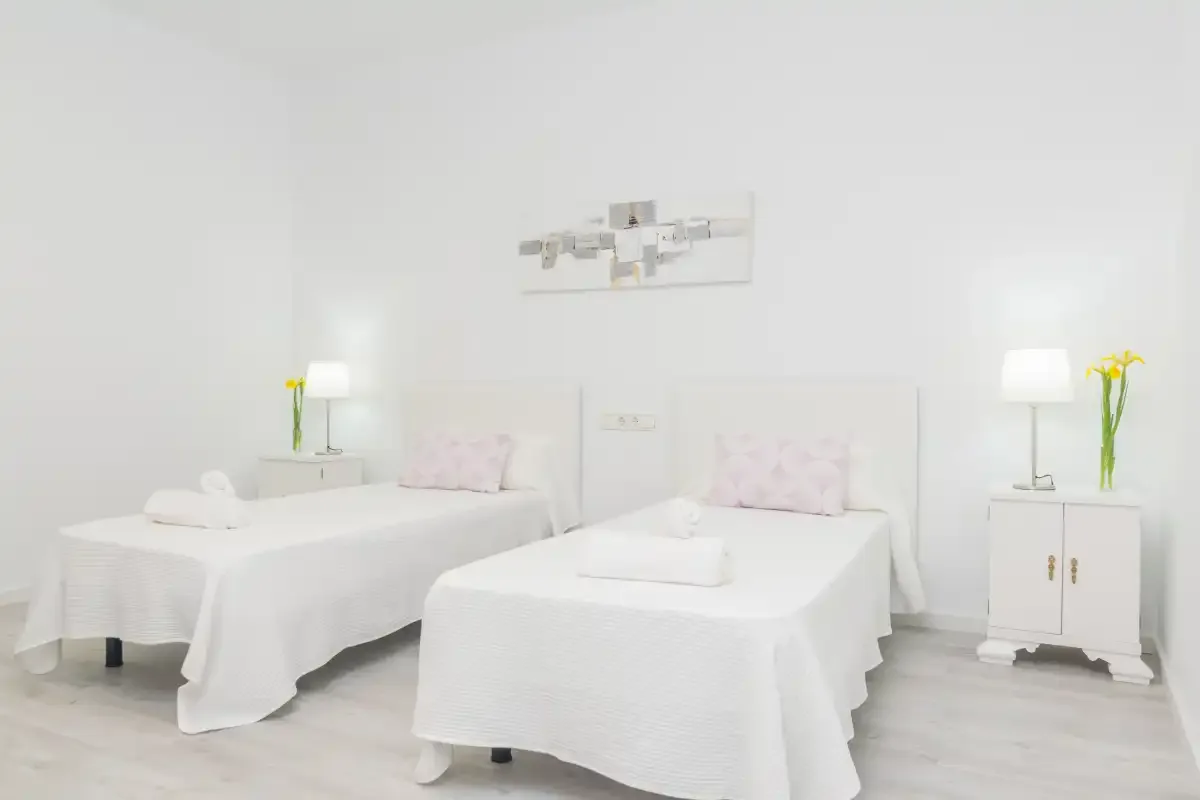 Un dormitorio luminoso tiene dos camas con ropa de cama blanca y mesitas a juego.