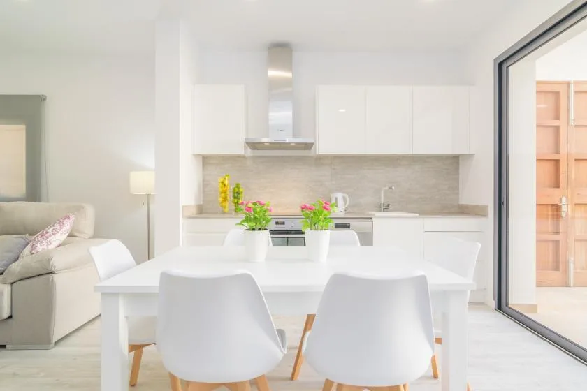 Un comedor luminoso con una mesa blanca y una cocina abierta al fondo.