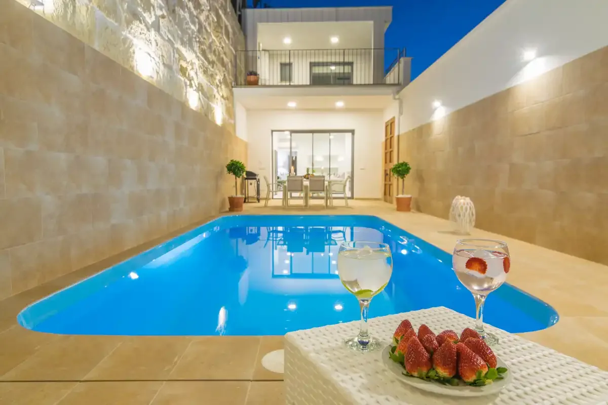 Una piscina en un patio frente a un edificio, con bebidas y fruta sobre una mesa.