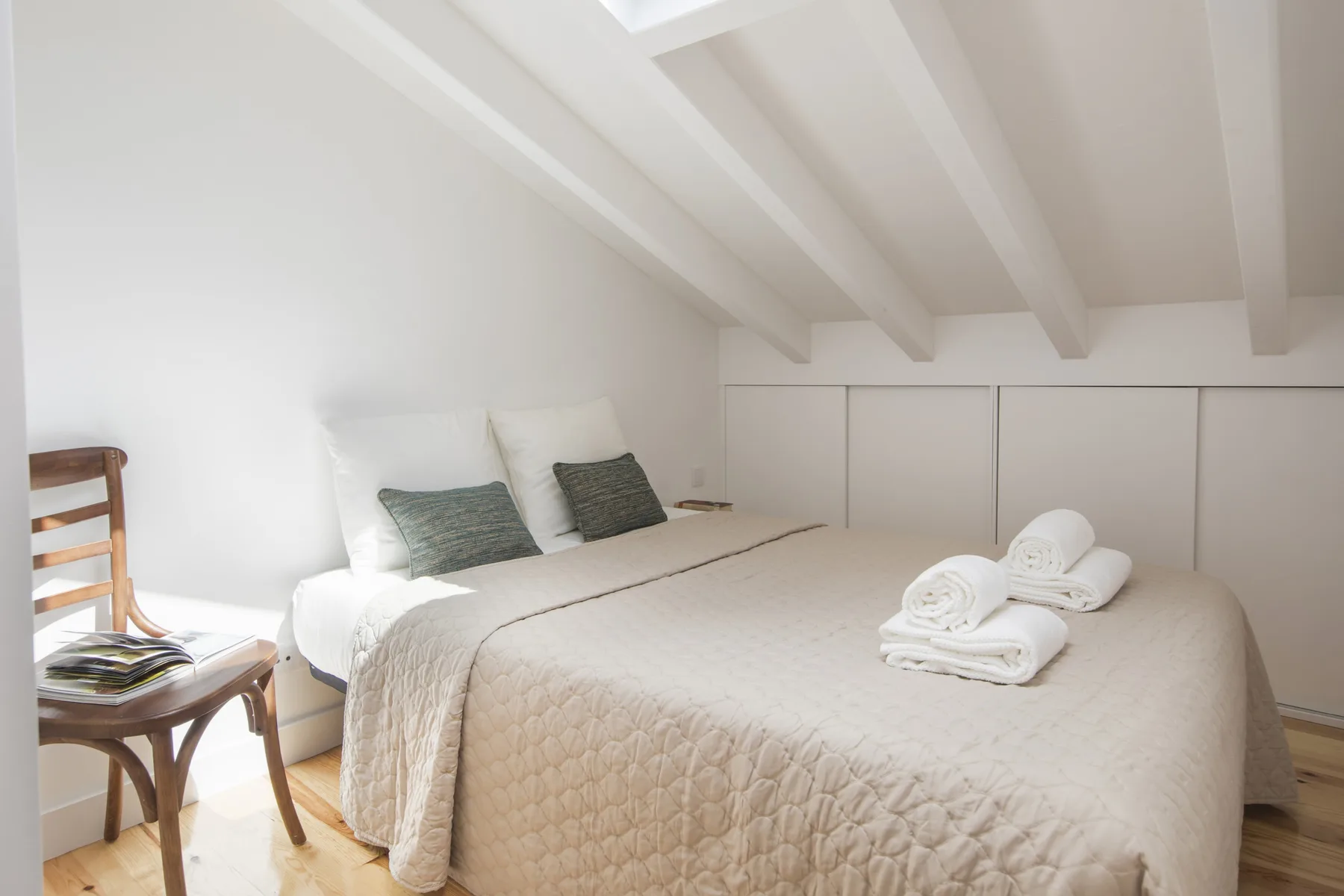 Dormitorio luminoso con una cama, toallas dobladas y techo inclinado.