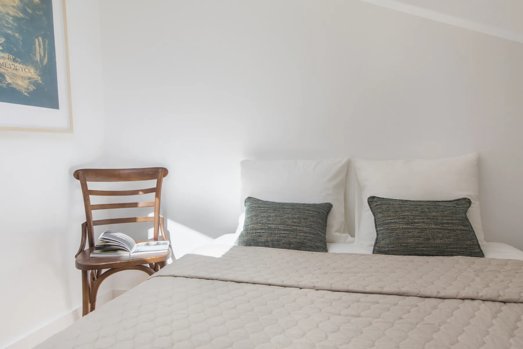 Un dormitorio luminoso con una cama bien tendida y una silla de madera al lado.