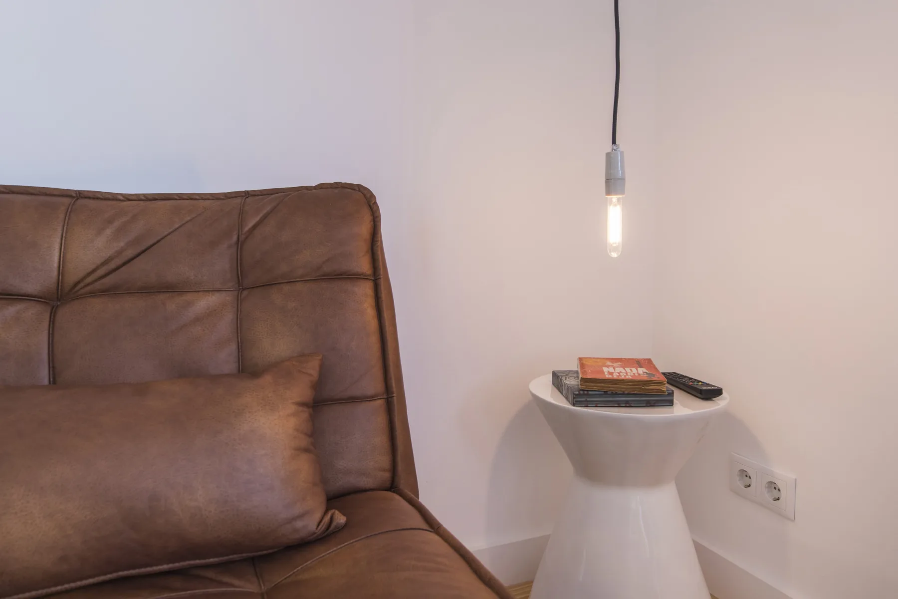 Una zona de descanso con un asiento tapizado marrón junto a una mesa pequeña y una lámpara colgante.