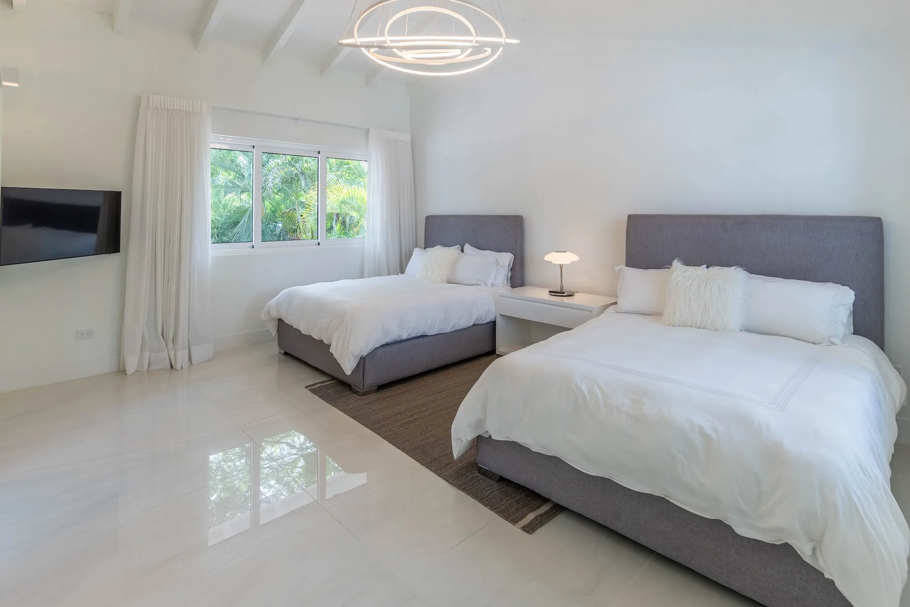 Un dormitorio luminoso con dos camas, una ventana y un televisor en la pared.