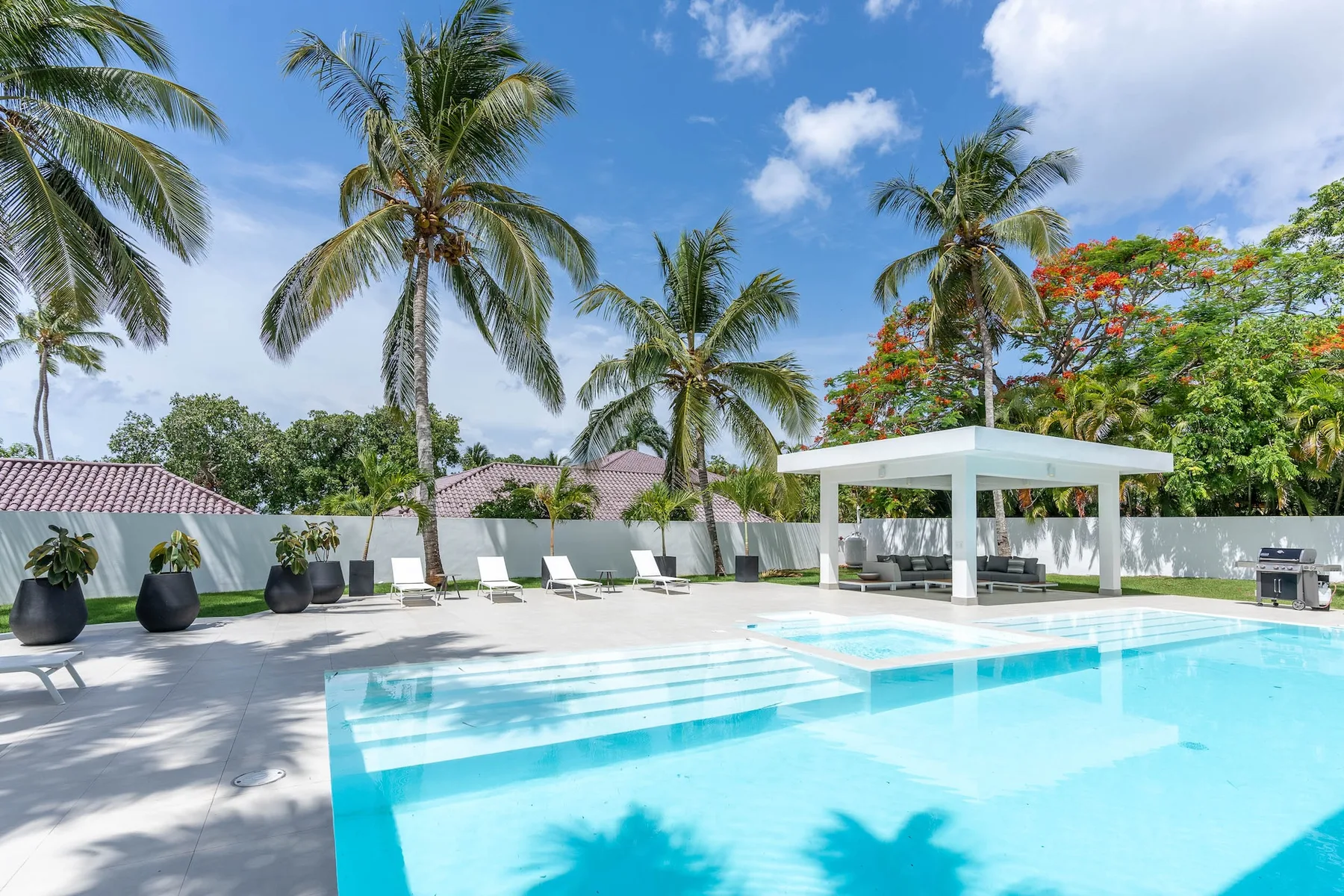 Una piscina exterior luminosa está junto a una zona de descanso sombreada y palmeras.