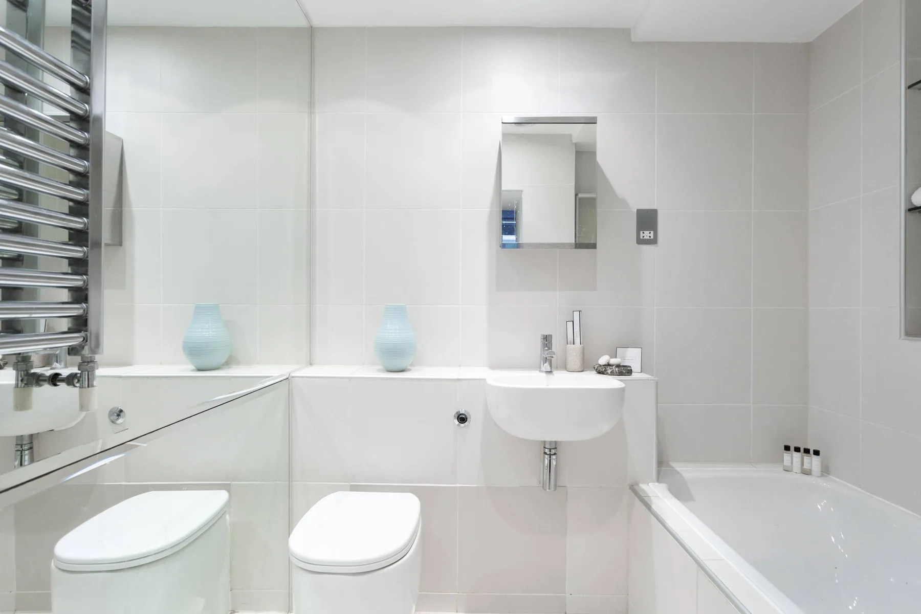 Un baño luminoso con azulejos, bañera, lavabo y dos inodoros.