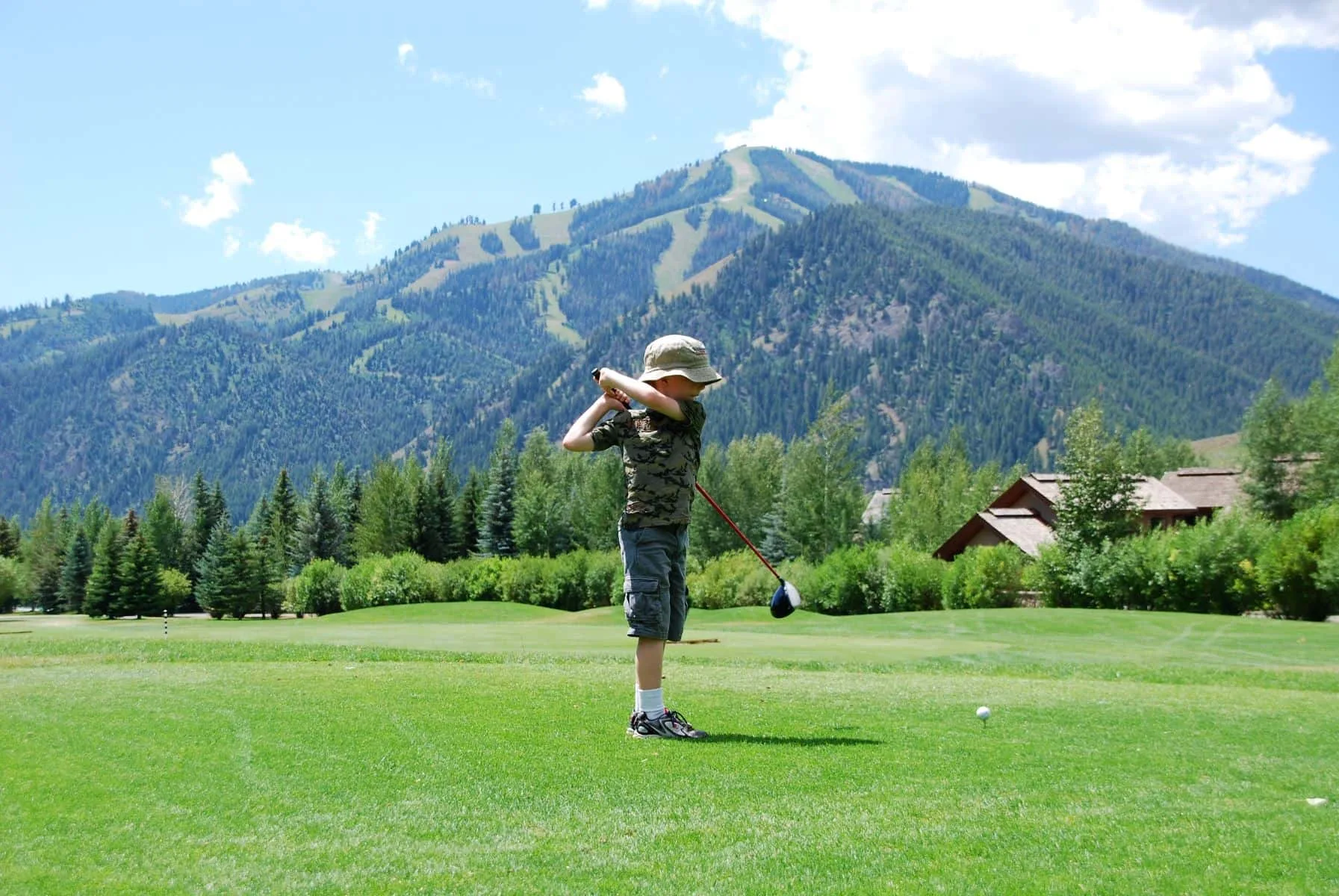 Una persona practica un swing de golf en un campo de césped, con una montaña al fondo.