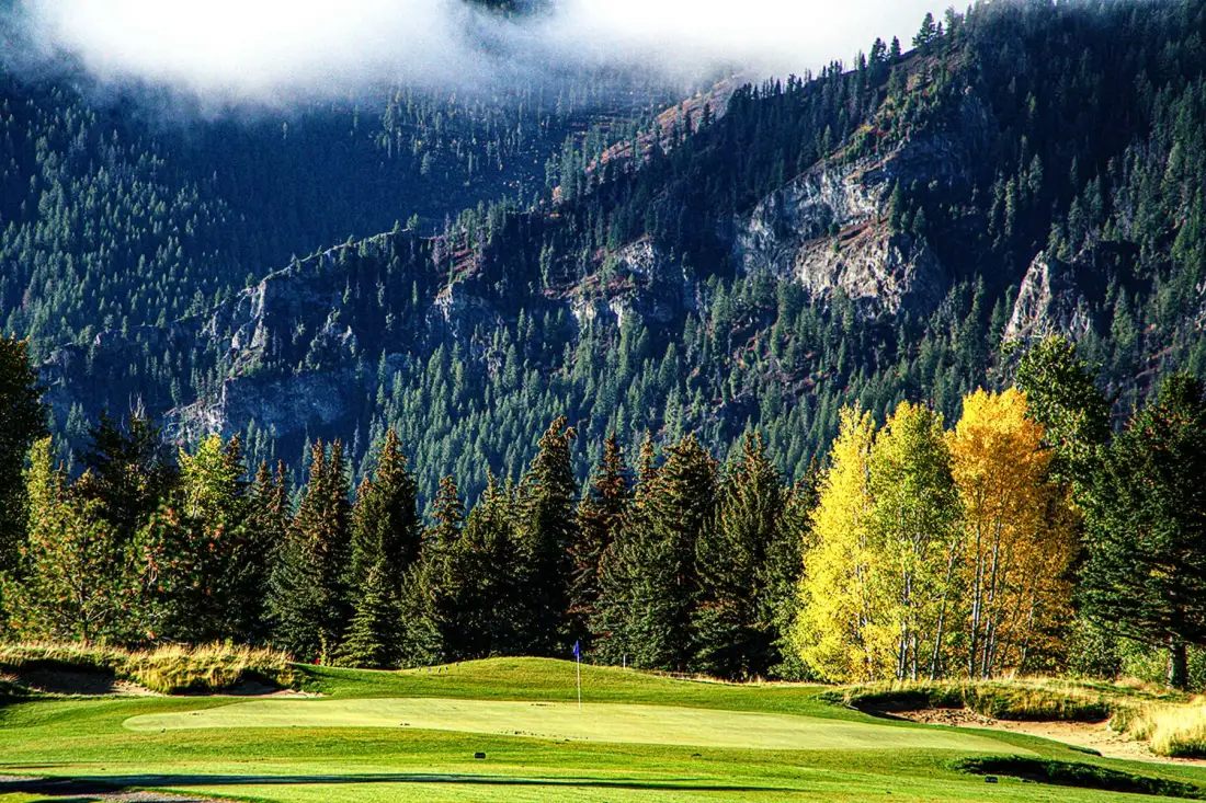 Un campo de golf bordea una ladera montañosa boscosa bajo nubes bajas.