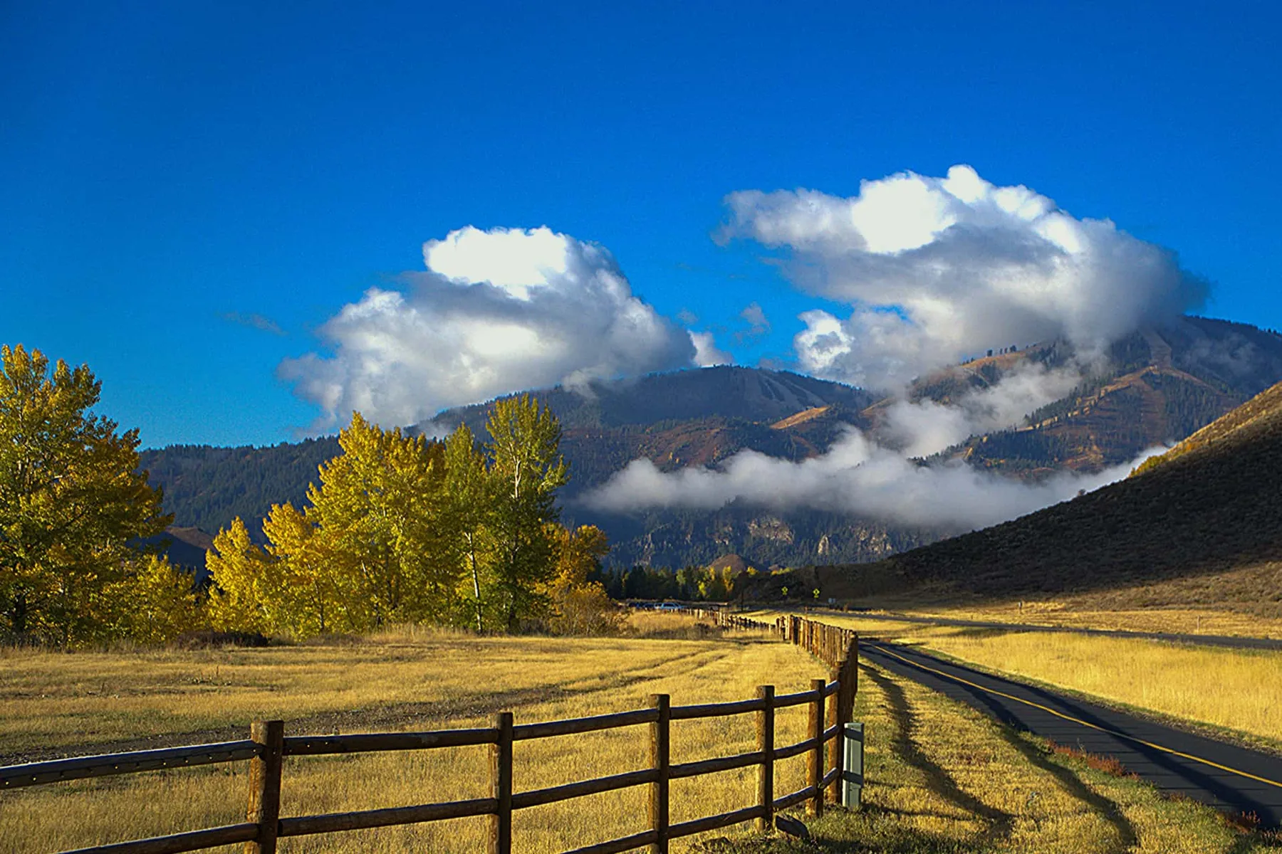 Una carretera rural bordea una cerca de madera en un campo dorado bajo montañas cubiertas de nubes.