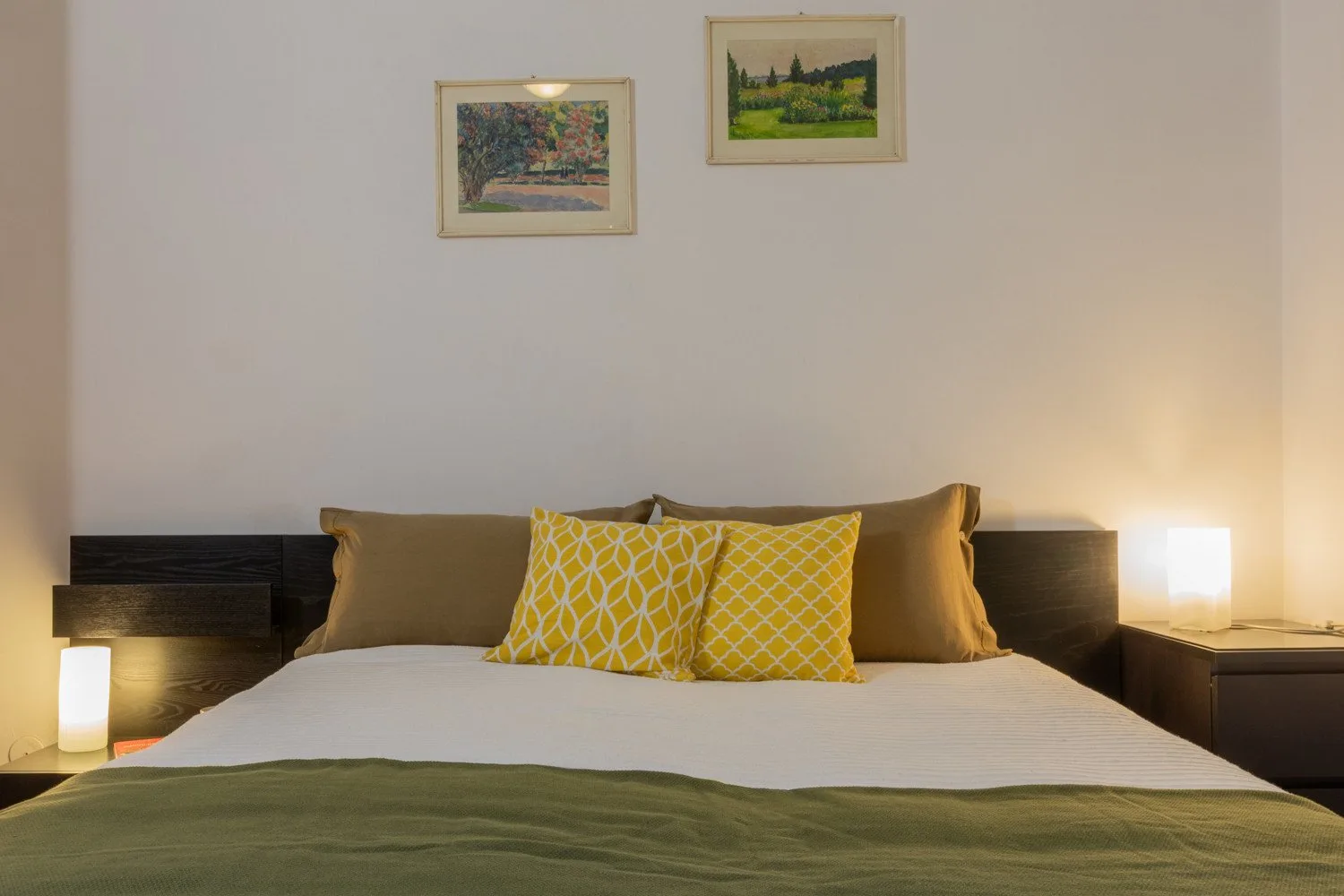A bedroom with a bed, yellow patterned cushions, and framed landscape pictures.