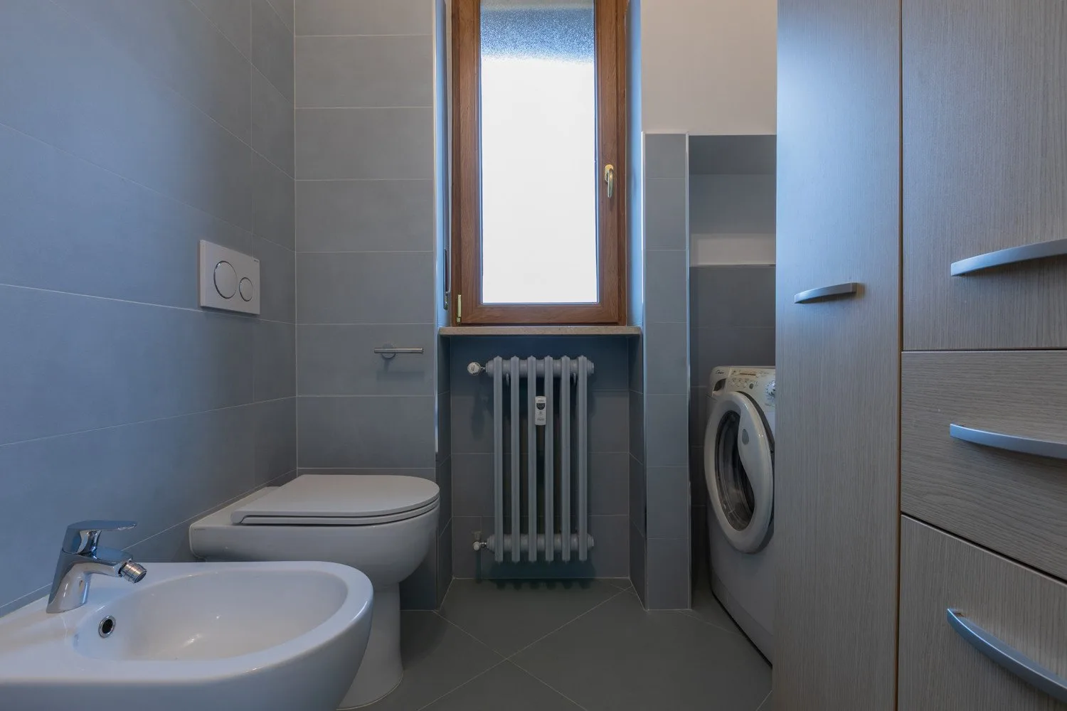A tiled bathroom with a sink, toilet, and window above a radiator.