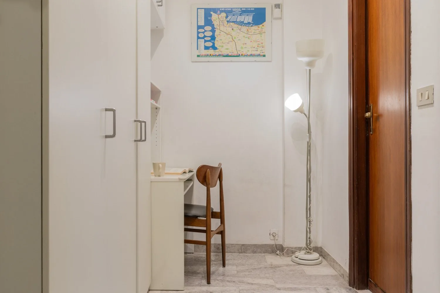 A small room with a desk, wooden seating, a floor lamp, and a closed door.