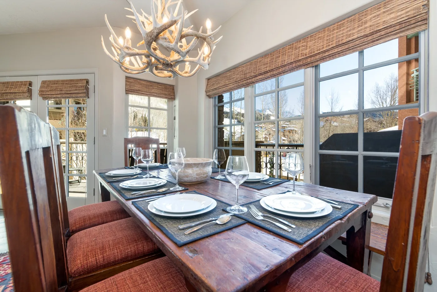 A dining area with a wooden table set with dishes beside large windows.