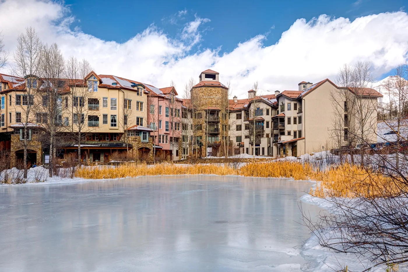 A multi-story building stands beside a frozen pond in a snowy outdoor scene.