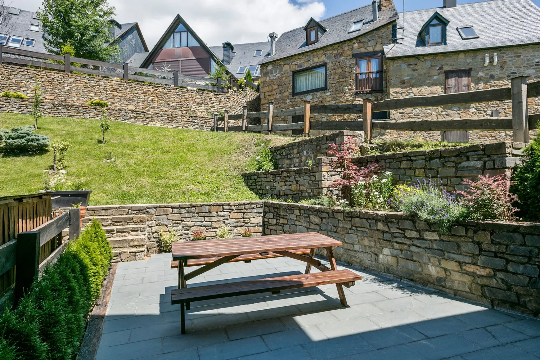 Outdoor patio area with a wooden picnic table set on a stone-tiled floor, surrounded by a garden with various plants, and houses with mountains in the background.