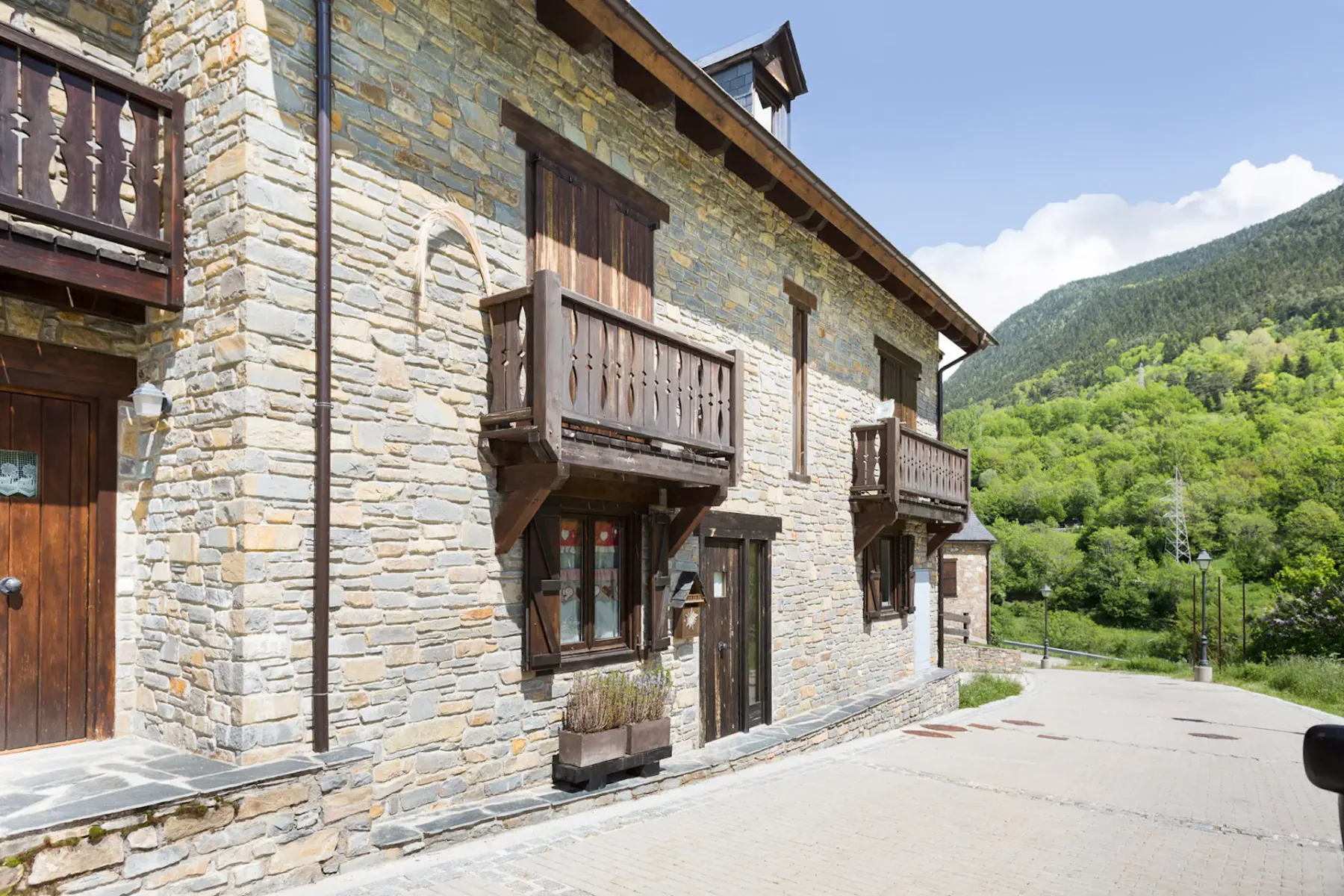 Stone building with wooden balconies and doors, situated on a paved roadway with green, forested hills and a blue sky in the background.