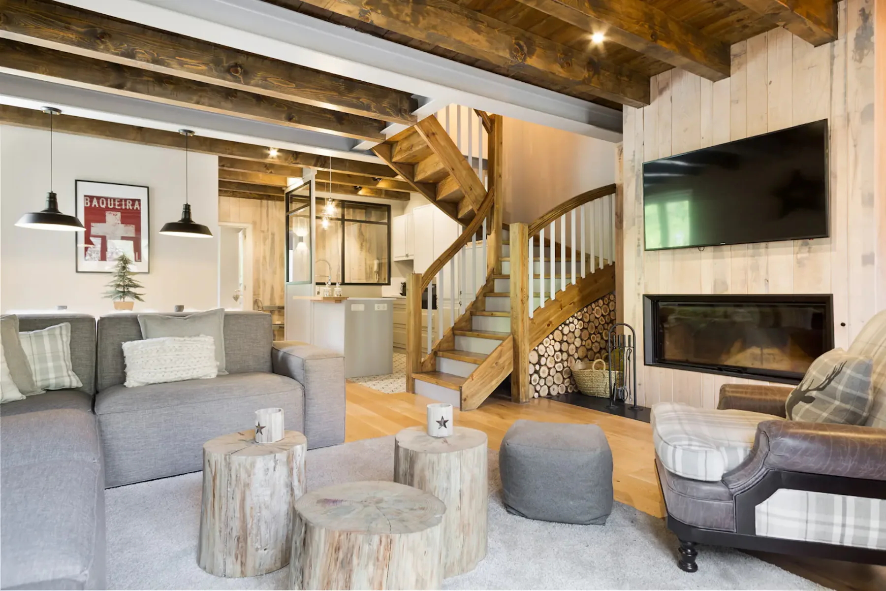 A living room with gray seating, round tables, and a wooden staircase beside a fireplace.