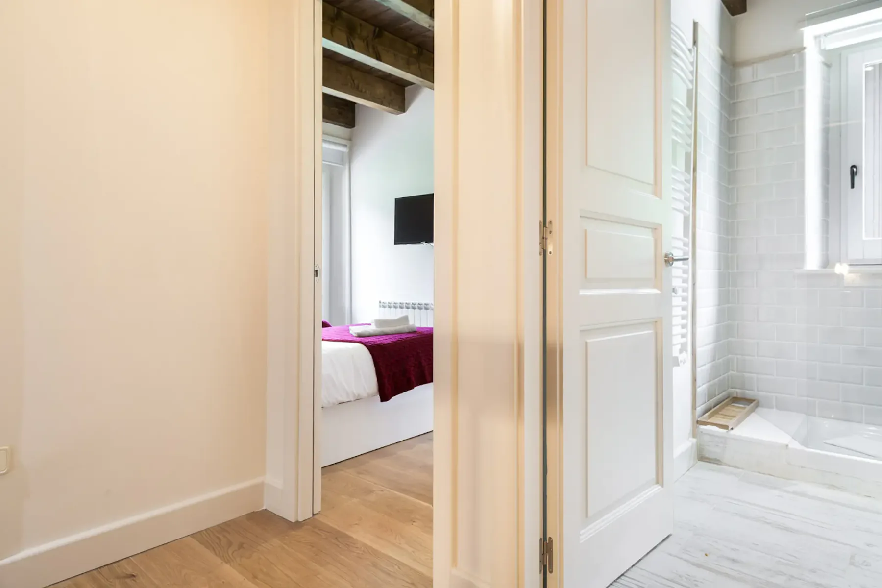 A bathroom entrance opens beside a bedroom with a bed and wall-mounted screen.