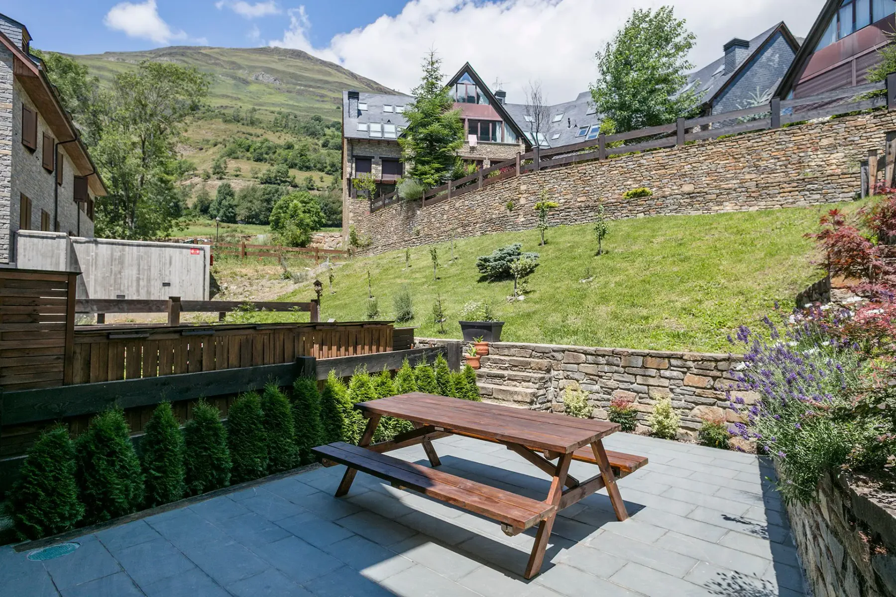 Outdoor patio area with a wooden picnic table set on a stone-tiled floor, surrounded by a garden with various plants, and houses with mountains in the background.