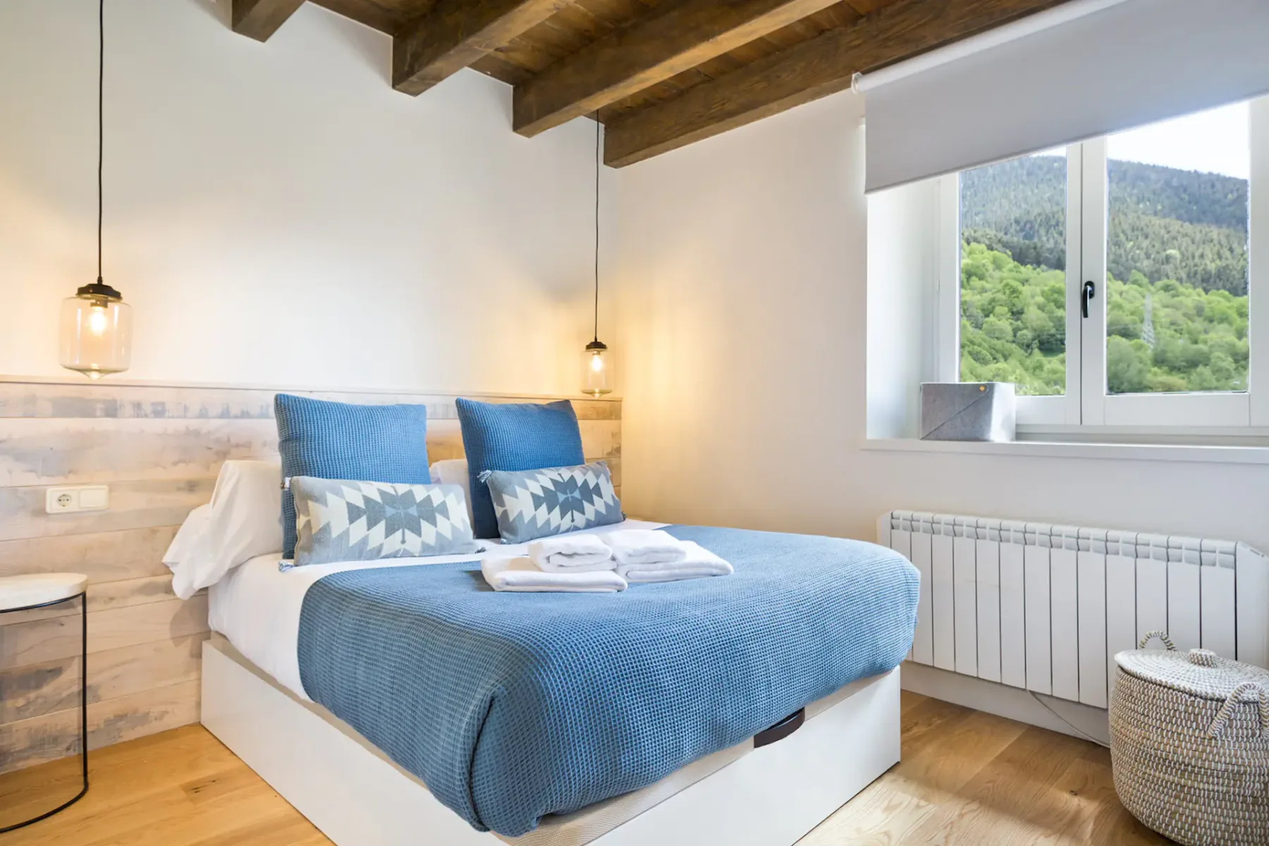 A bedroom with a blue bed and a window overlooking green hills.