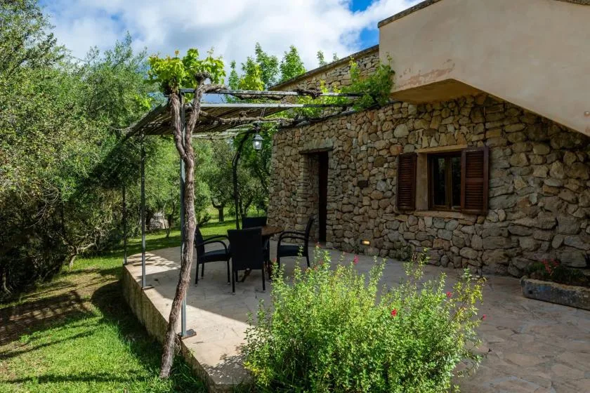 A stone building opens onto a paved patio with outdoor seating and a leafy pergola.