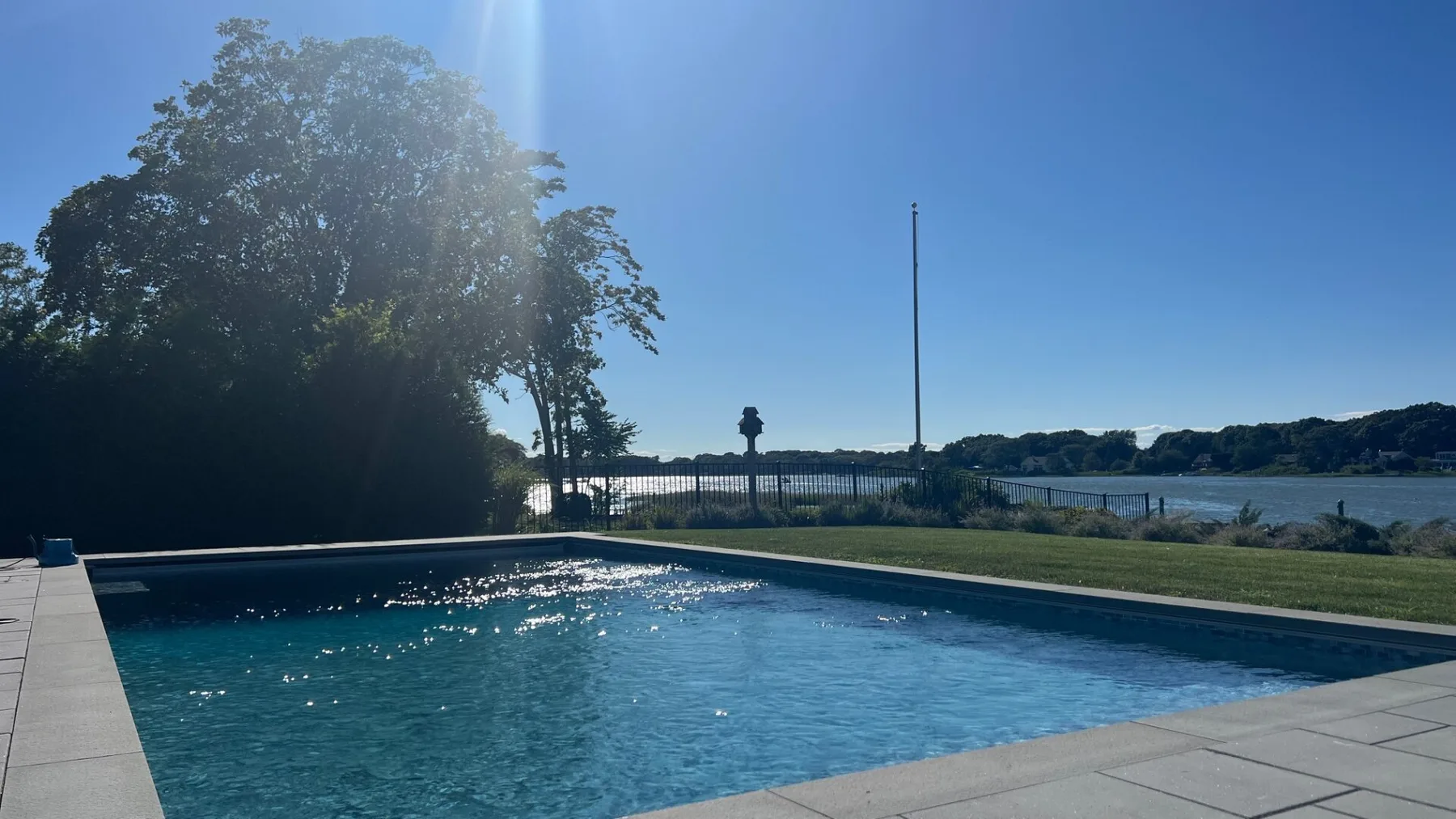 An outdoor swimming pool sits beside a paved deck, with water visible beyond.
