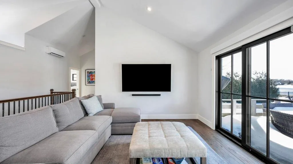 Una sala luminosa tiene una zona de asientos, un televisor en la pared y grandes puertas de vidrio.