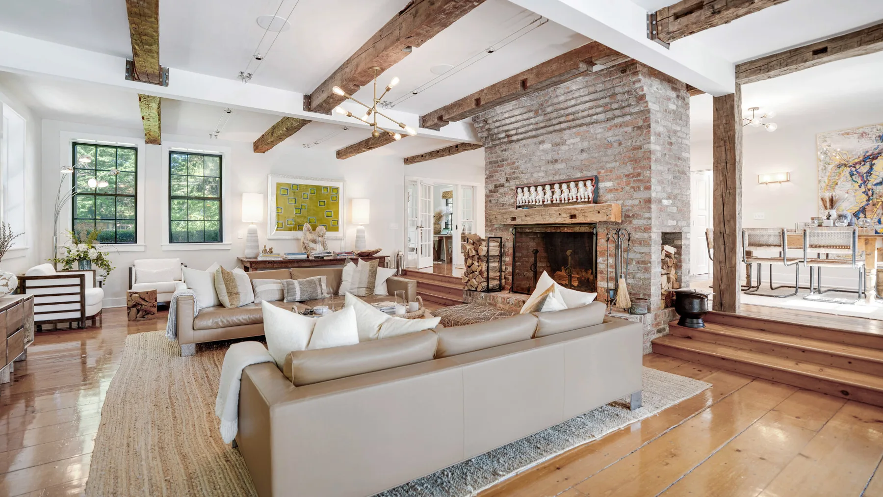 A spacious living room features seating around a brick fireplace and exposed wooden beams.