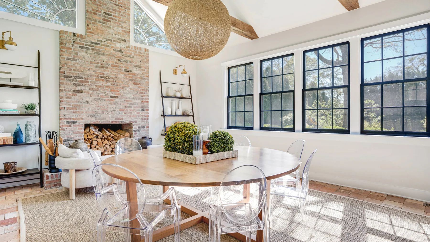 A bright dining area with a large table beside tall windows and a brick fireplace.