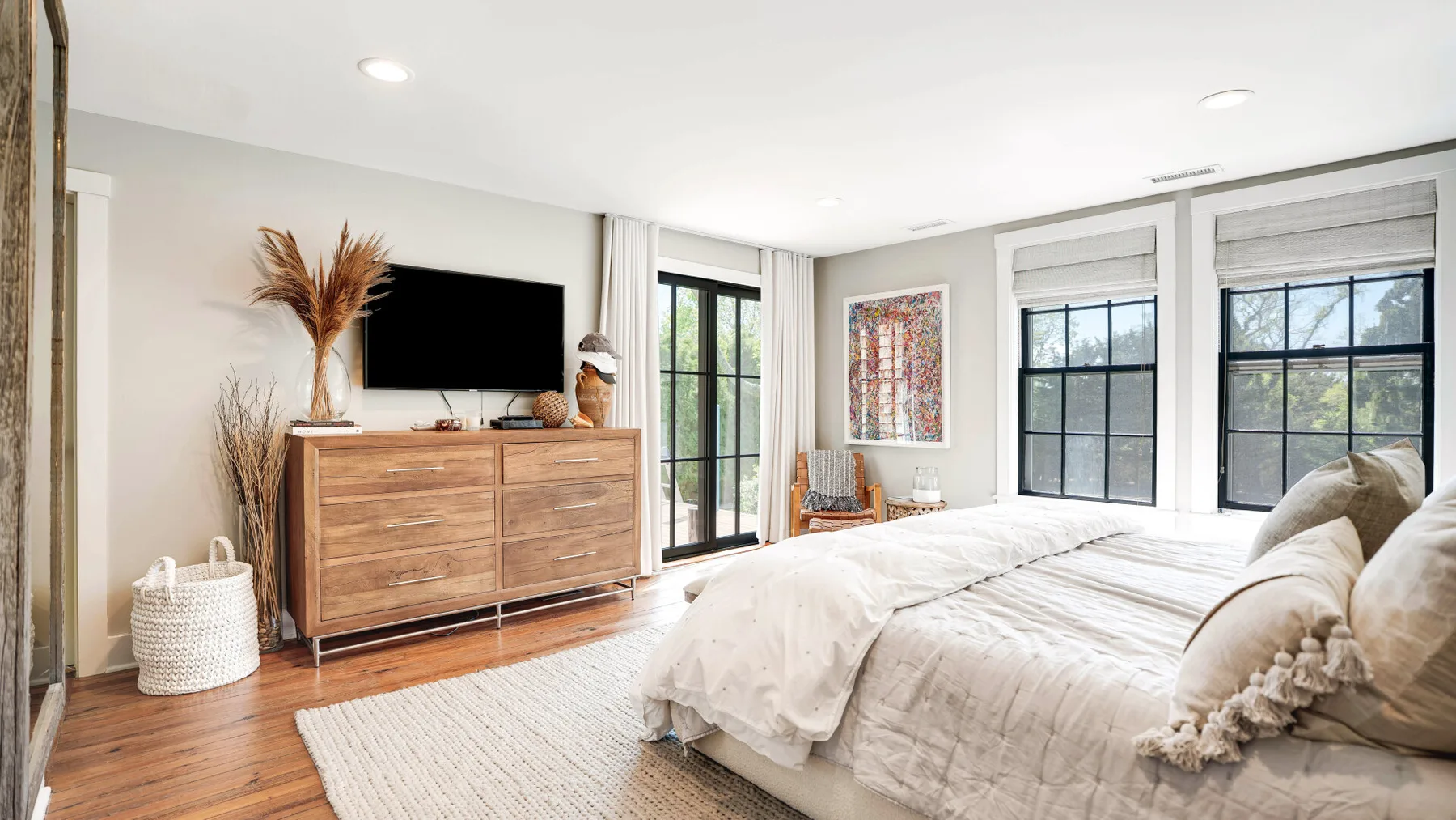 A bright bedroom with a large bed, wooden dresser, and tall windows.