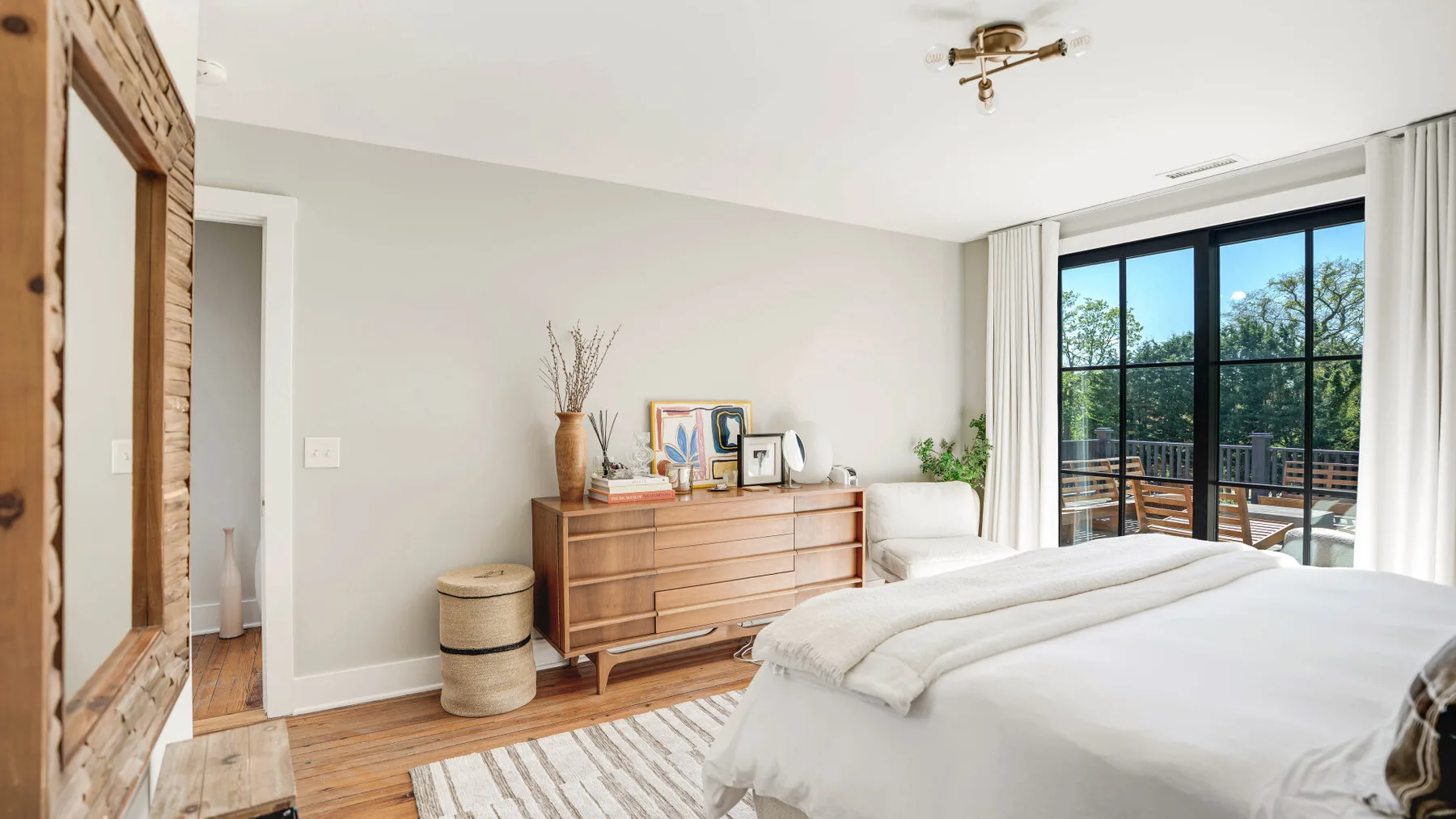 A bright bedroom with a large bed, wooden dresser, and glass doors opening to a balcony.