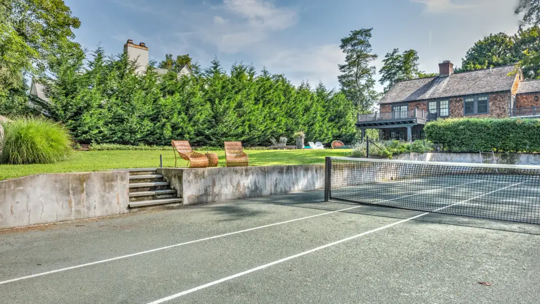 Una pista de tenis exterior está junto a un jardín cuidado y una casa de ladrillo.