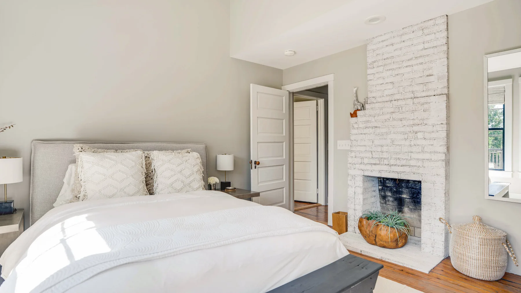A bright bedroom with a neatly made bed beside a white brick fireplace.