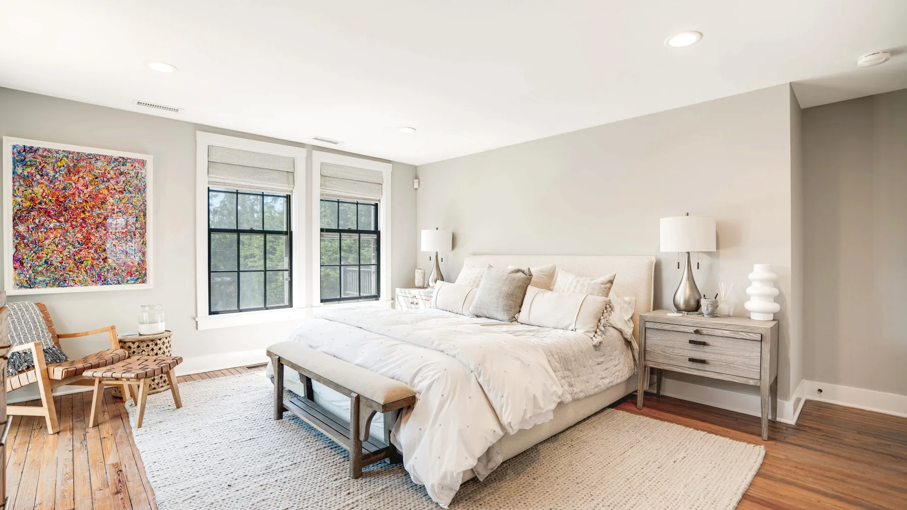 A bright bedroom features a large bed, bedside tables, and colorful wall art.