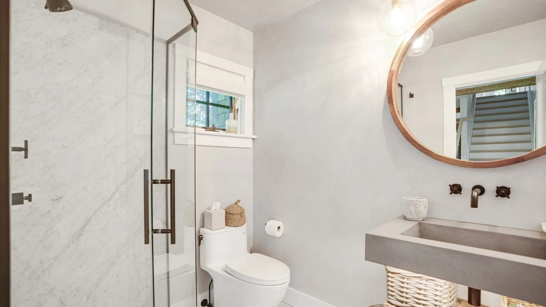 A bright bathroom with a glass shower, toilet, and sink beneath a round mirror.