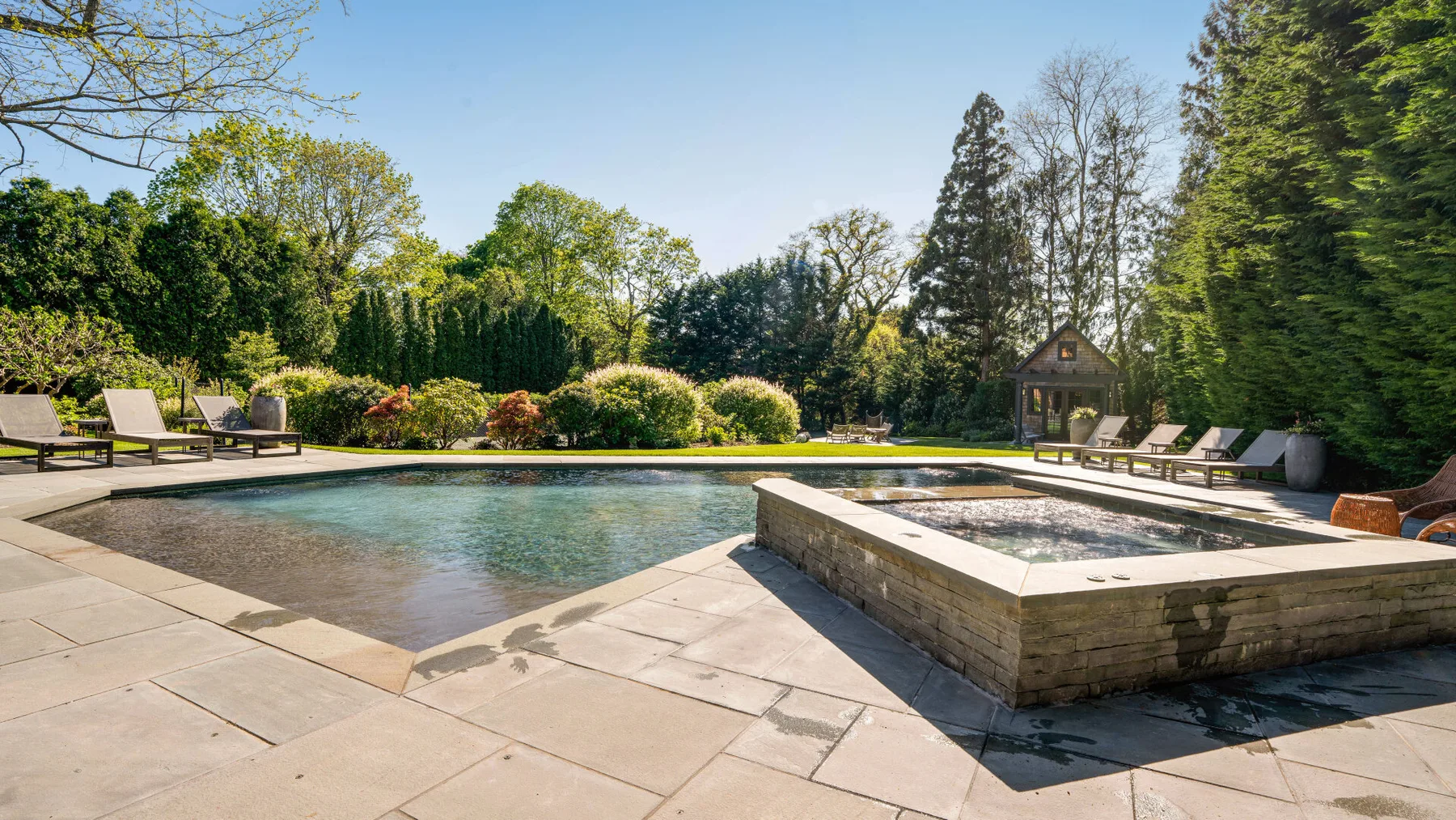 A garden pool with a raised square spa and lounge seating on a sunny patio.