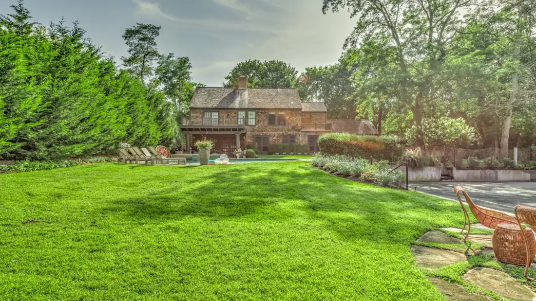A large house stands beyond a green lawn, with trees and outdoor seating.