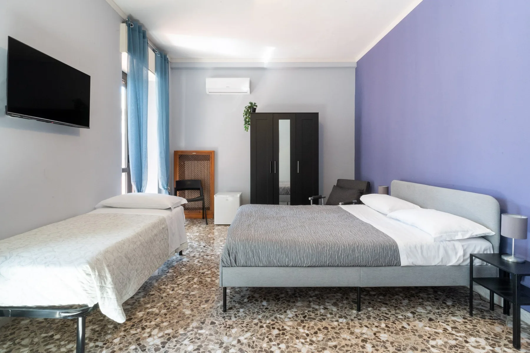Un dormitorio con dos camas, un armario y ventanas altas con cortinas azules.