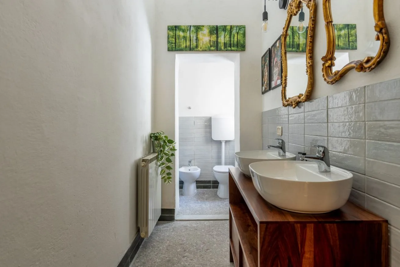 A bathroom with a wooden vanity, vessel sinks, and a toilet in the adjoining area.