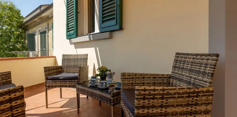 A sunny terrace with woven seating beside a cream-colored wall and open shutters.